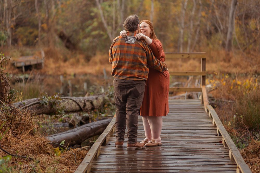 A couple stands on a rustic wooden boardwalk surrounded by autumn foliage during a romantic photoshoot.