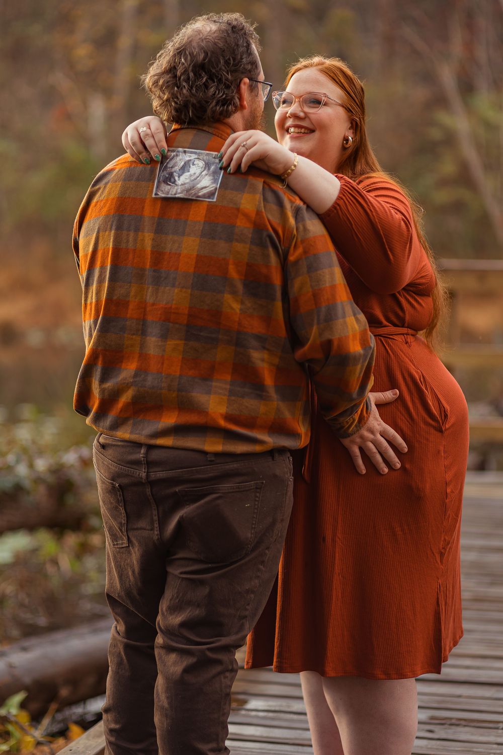 A couple in autumn attire share an intimate moment while walking on a wooden boardwalk through nature.