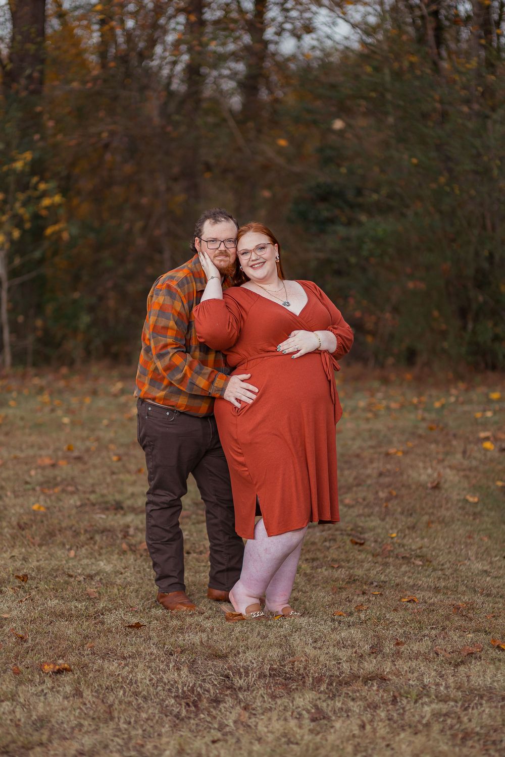 A couple poses outdoors in autumn, with one wearing a plaid shirt and the other in a red dress showing a baby bump.