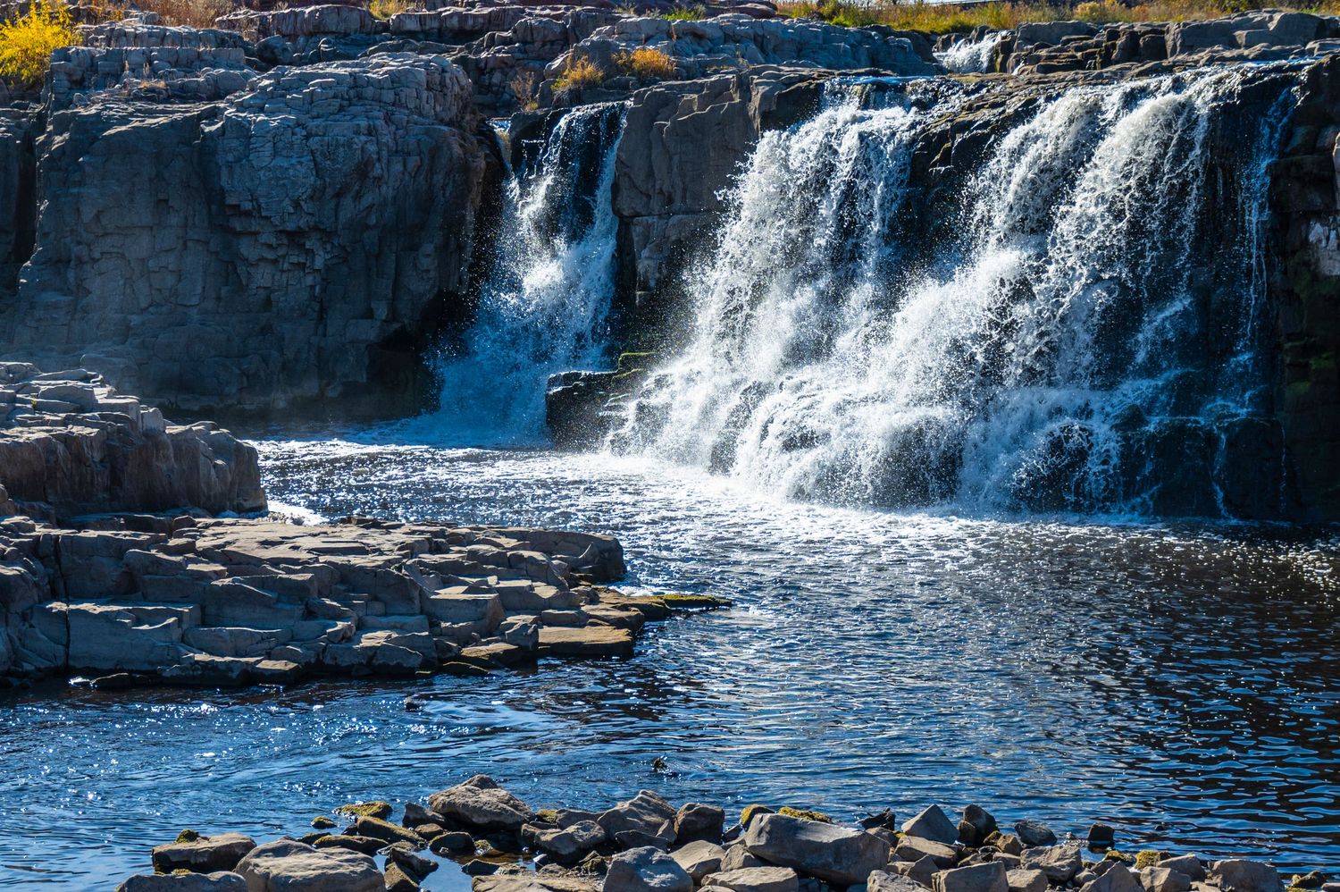 Wide waterfall cascades over dark rocky cliffs into a blue pool surrounded by stones.