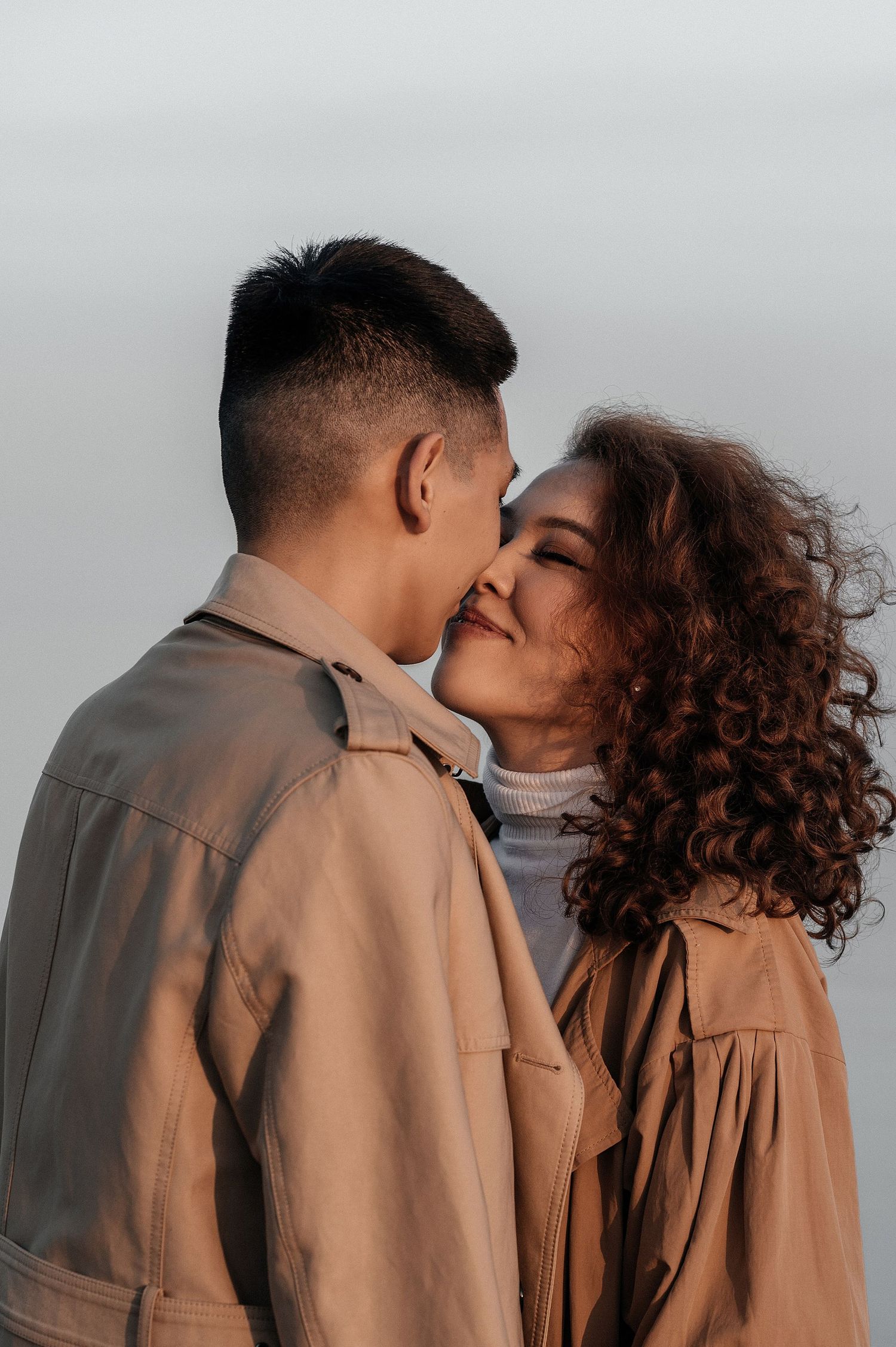 A couple in matching trench coats smiling at each other in the light of the sunset