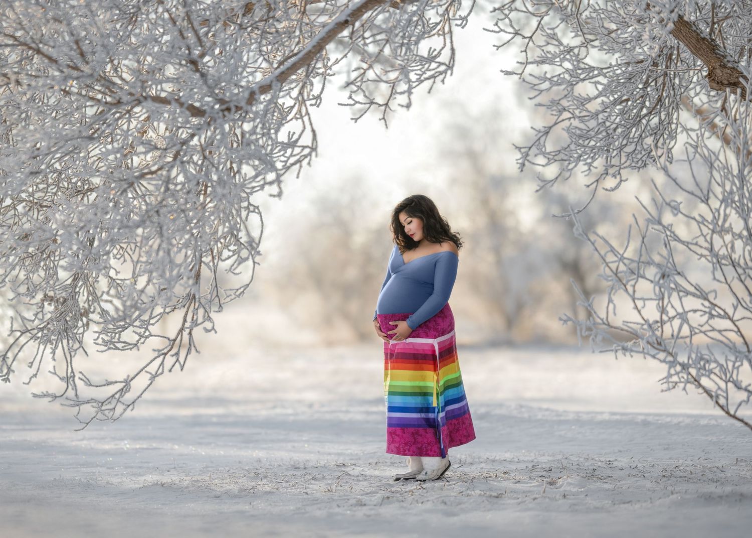 Maternity portrait featuring a lady in an Indigenous rainbow ribbon skirt on a winter landscape with frosted trees in Saskatchewan, Canada