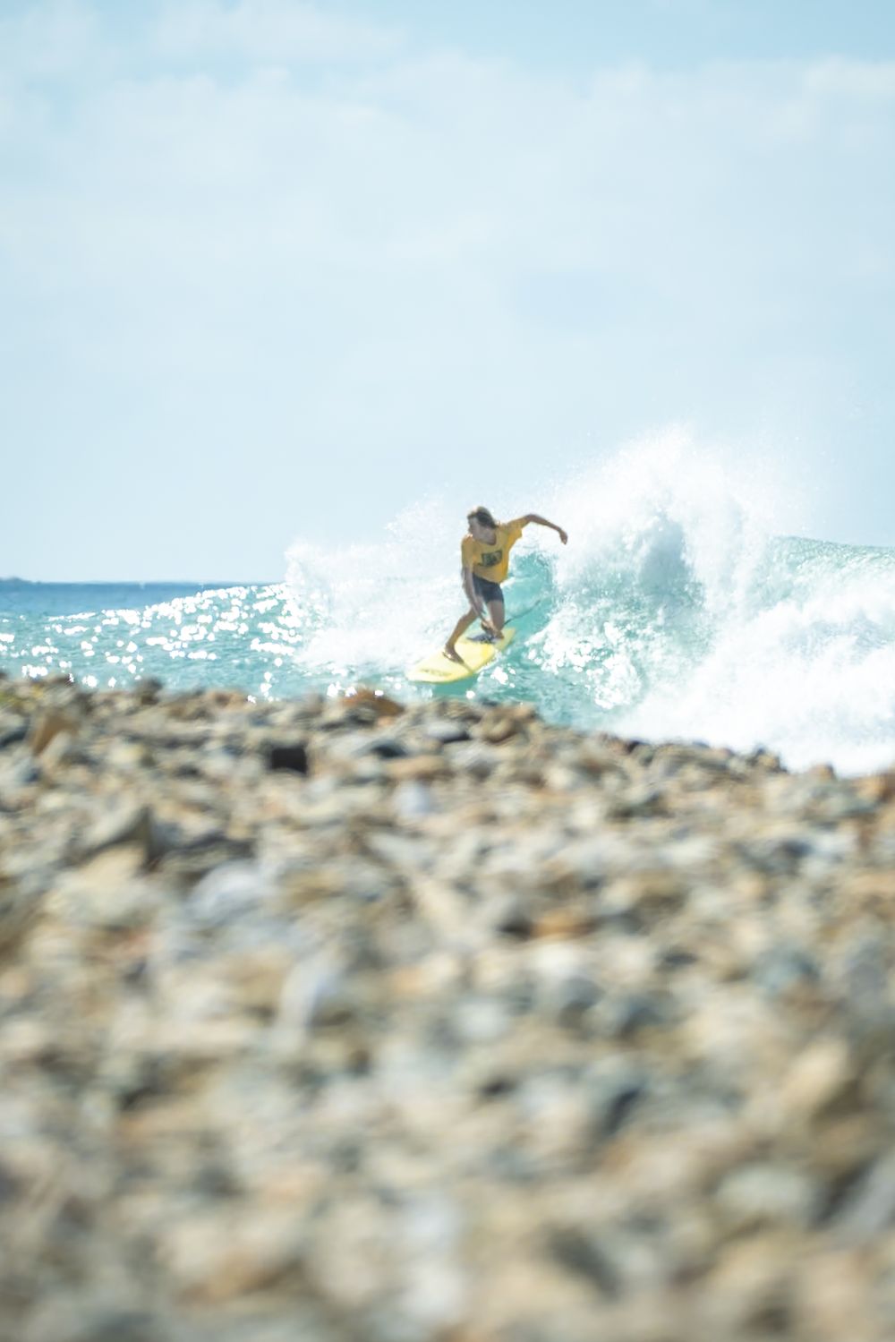 Surfer silhouetted against ocean spray with rocky shoreline in foreground.