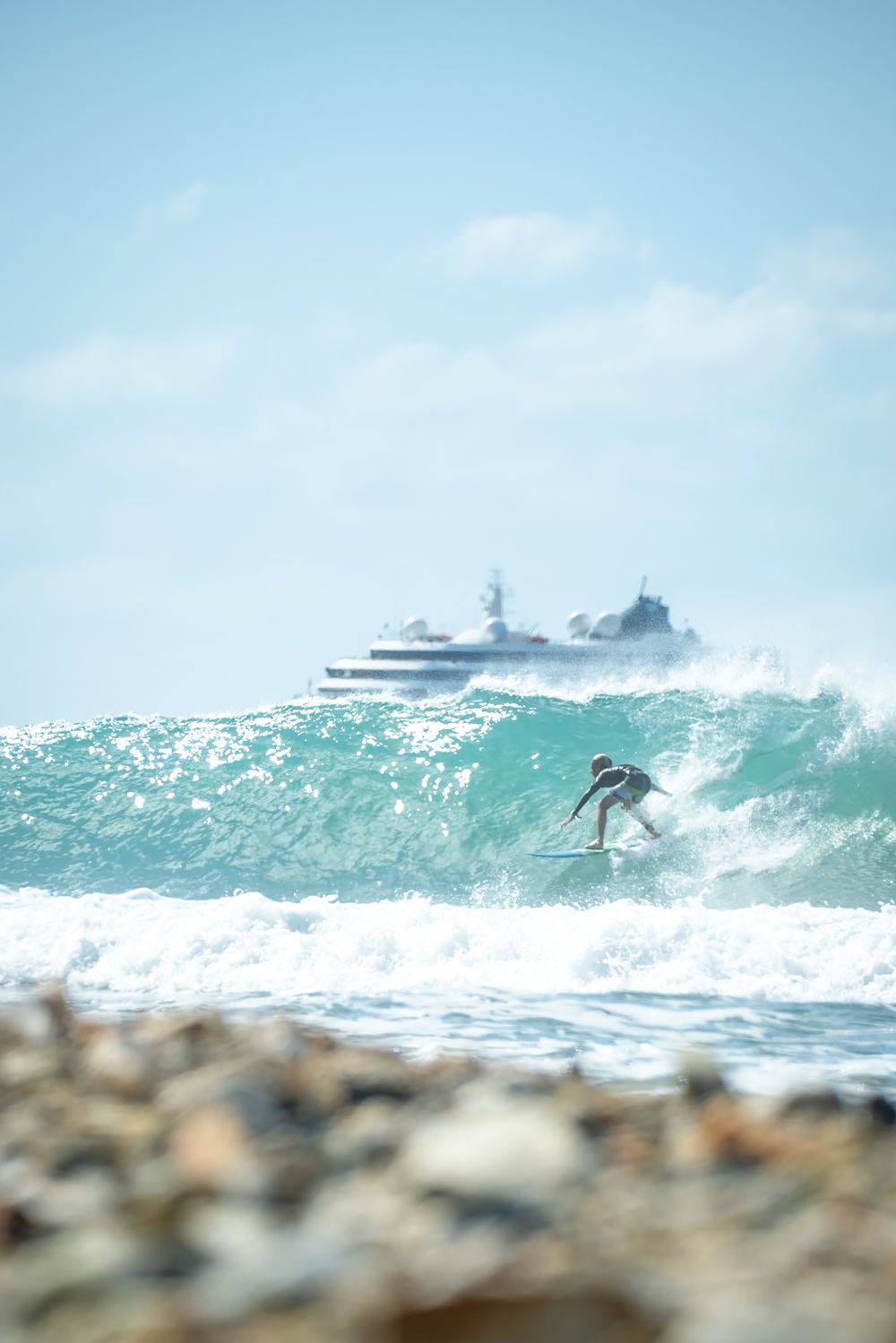 Surfer catching wave with luxury yacht visible in background on tropical waters.