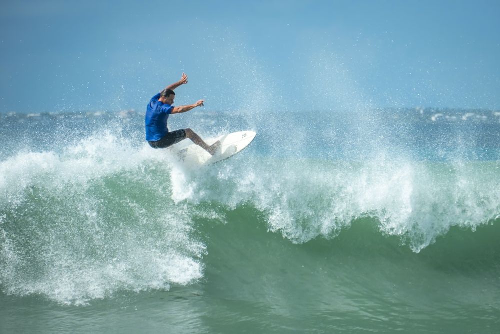 Surfer in blue shirt catching air off breaking wave during sunny day.