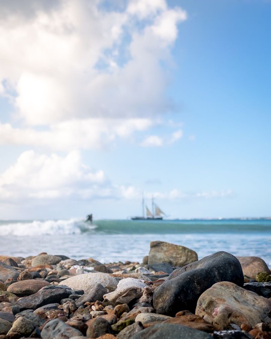 Rocky beach shoreline with sailing ship visible on horizon and crashing waves on tropical waters.