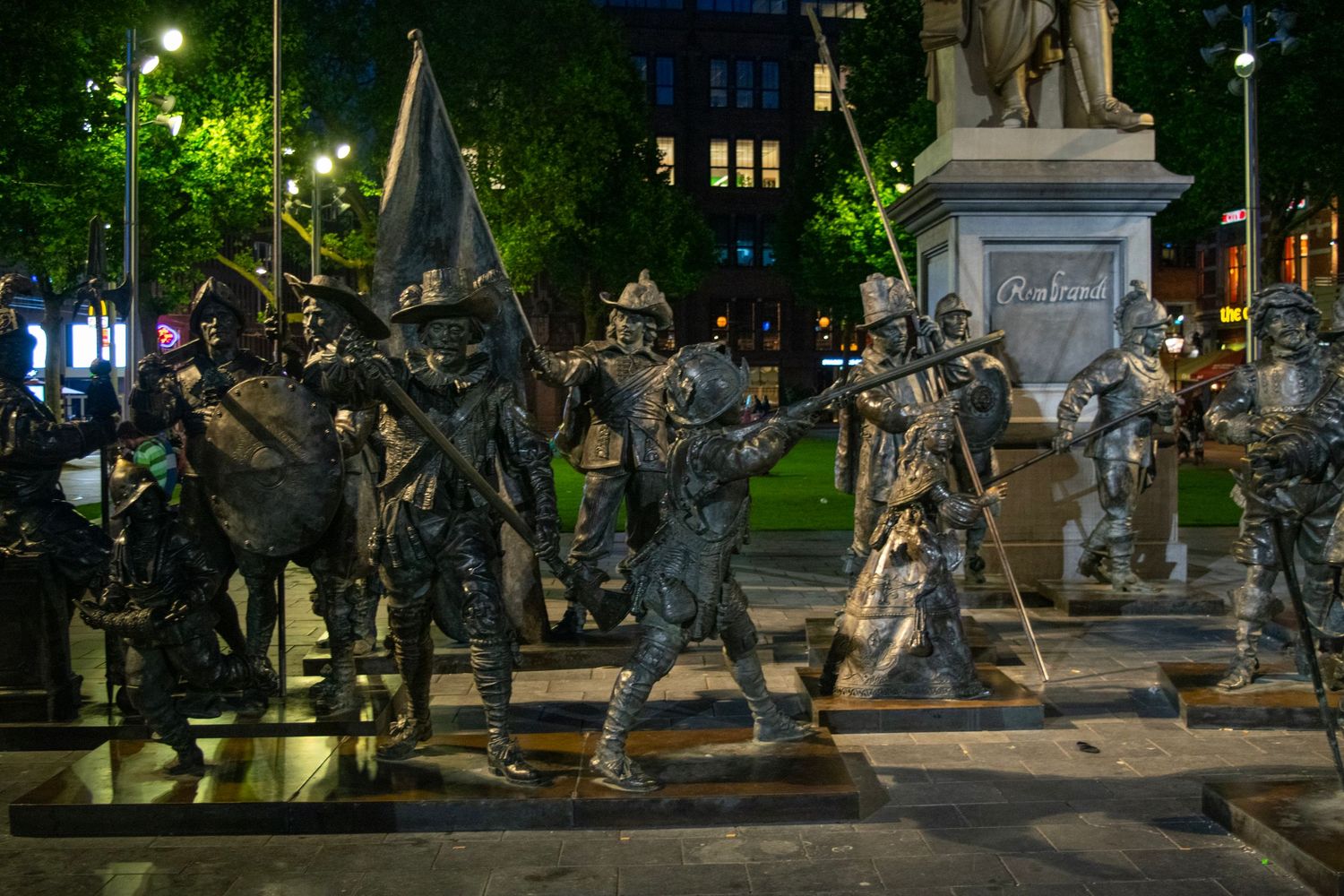 Metal statues stand illuminated at night in an urban plaza.