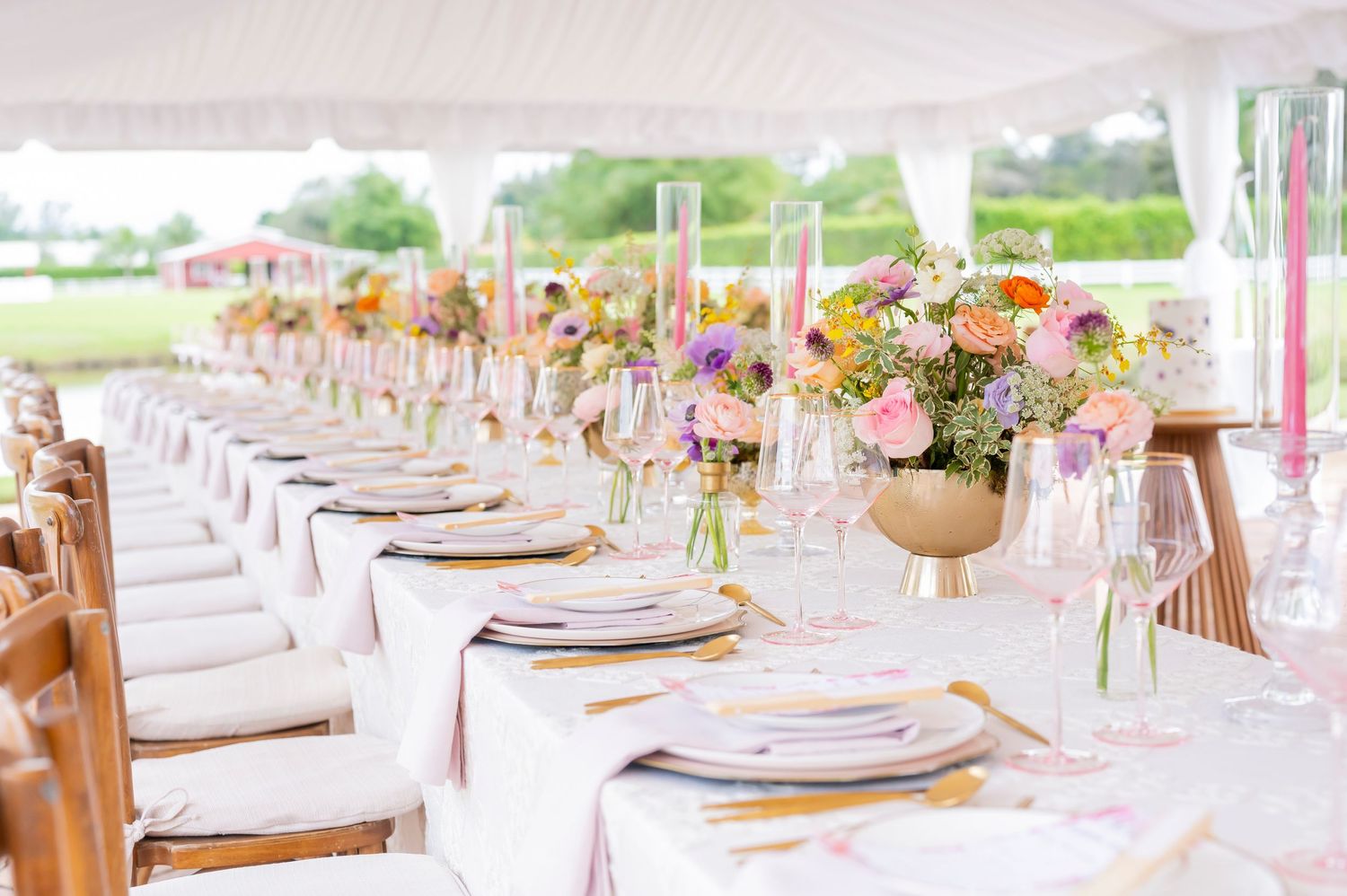 Long elegant reception table set with pink floral centerpieces, gold plates and natural wood chairs under white tent.