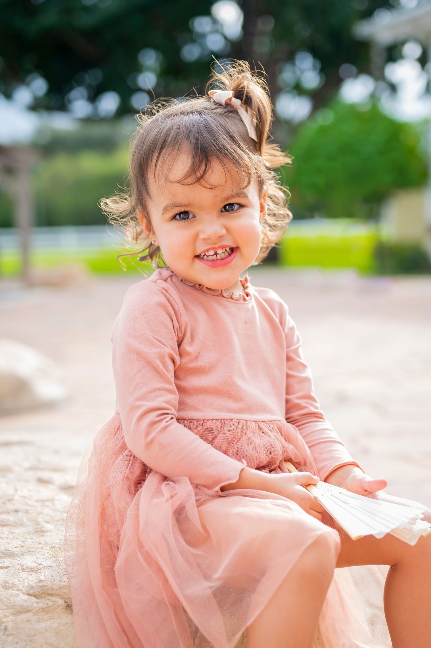 Toddler wearing pink dress smiles brightly while sitting outdoors on sunny day.