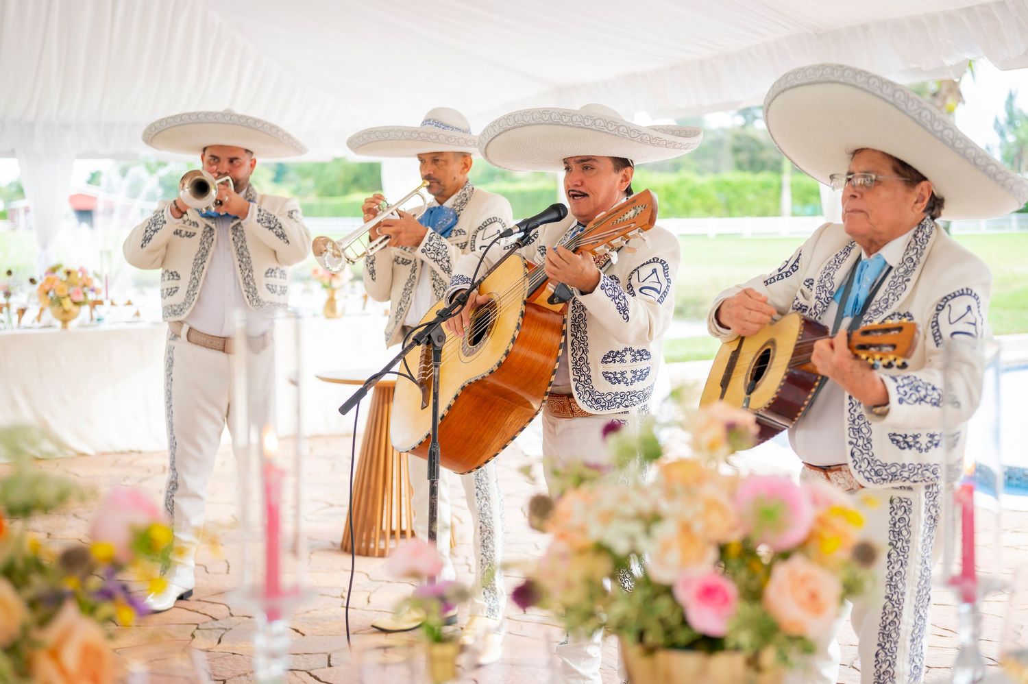 Traditional mariachi band in white and silver embroidered suits perform under a tent with pink floral decorations.