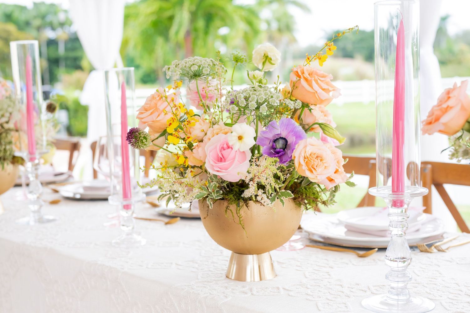 Elegant table setting with pink taper candles and a gold vase filled with colorful spring flowers on white linen tablecloth.