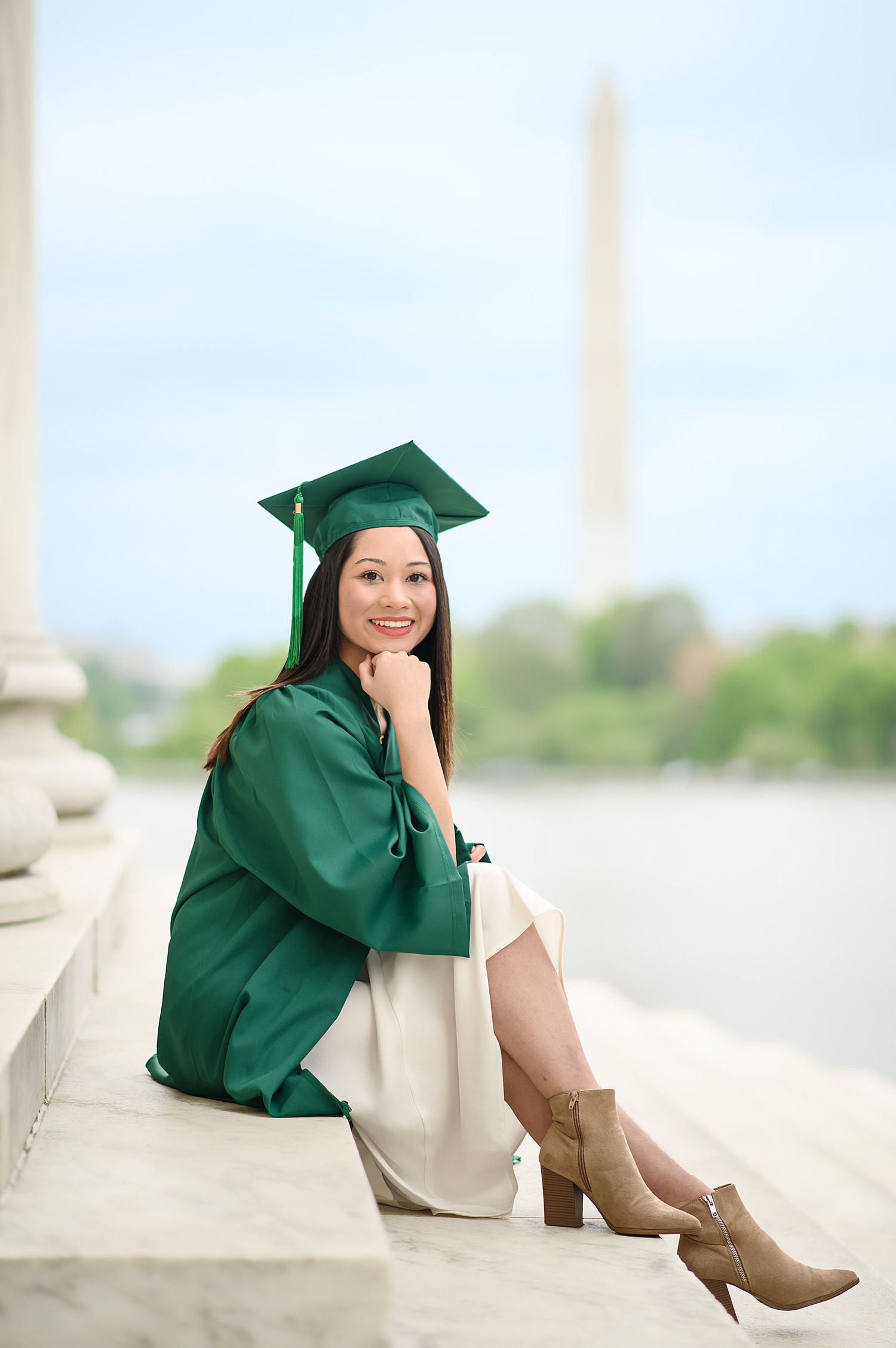 Graduate in emerald cap and gown poses on steps with brown ankle boots.