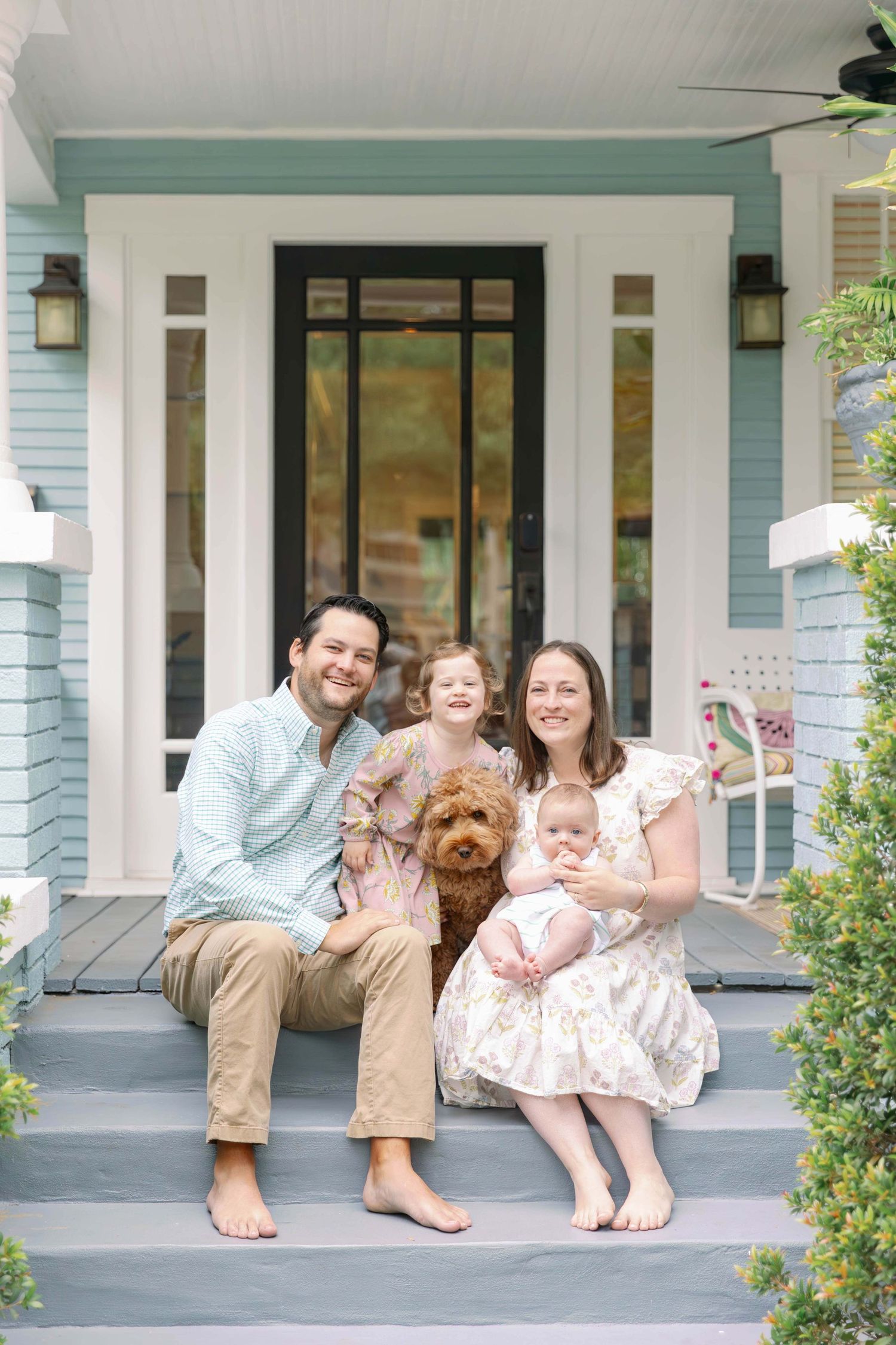 Family stands in front of house with American flag displaying twin babies.