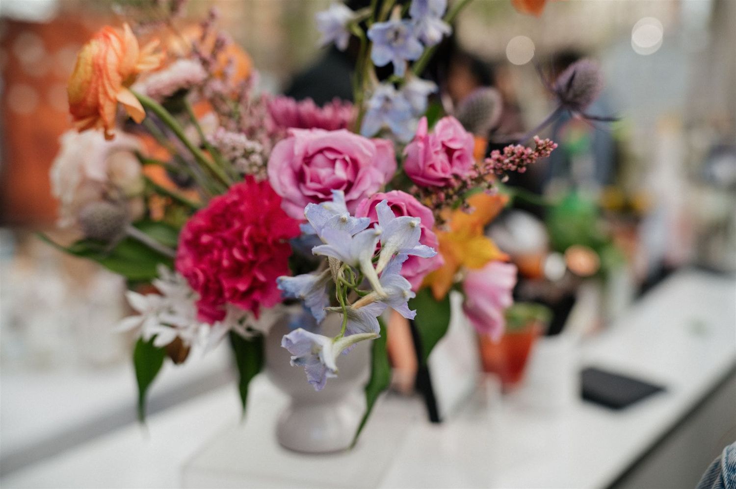 A vibrant floral arrangement with pink peonies, purple delphinium, and mixed blooms in a pale ceramic vase.