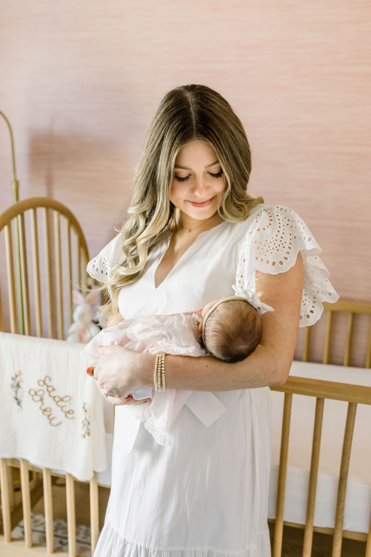 Parent in white lace dress holding baby in nursery with wooden crib.