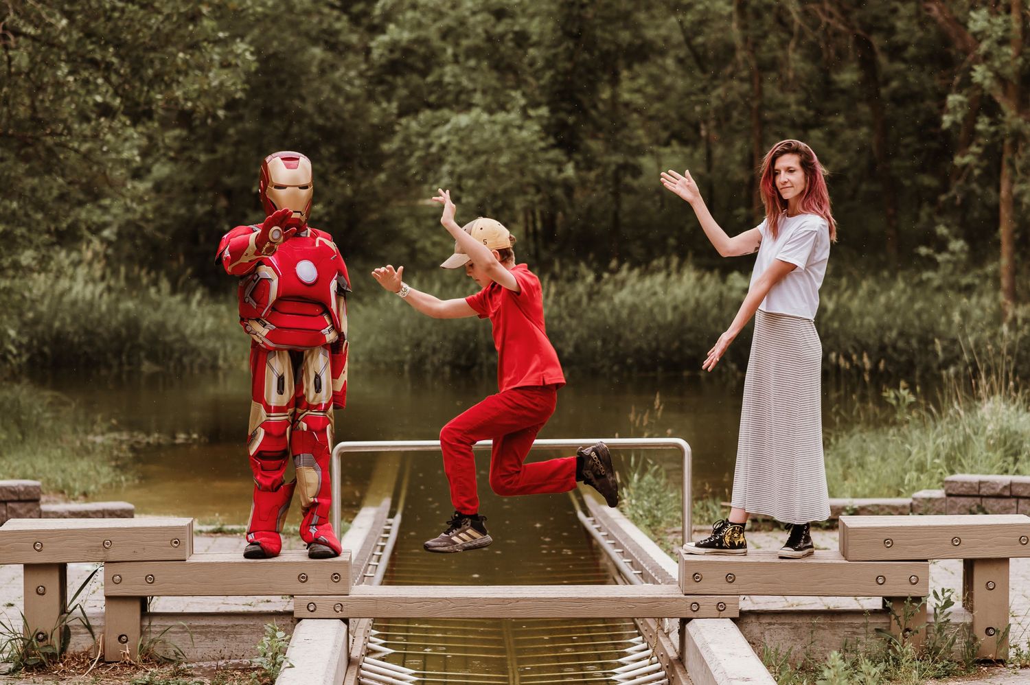 Three people stand and jump playfully on a wooden dock over a pond on a sunny summer day.