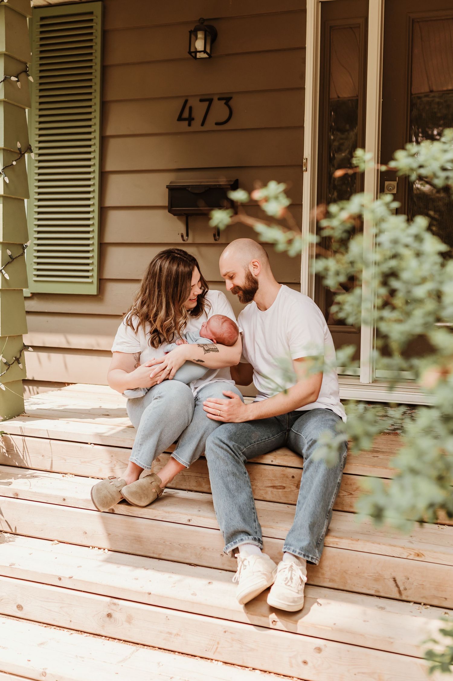 A family sits together on wooden porch steps outside a house with white siding and flowers in the foreground.