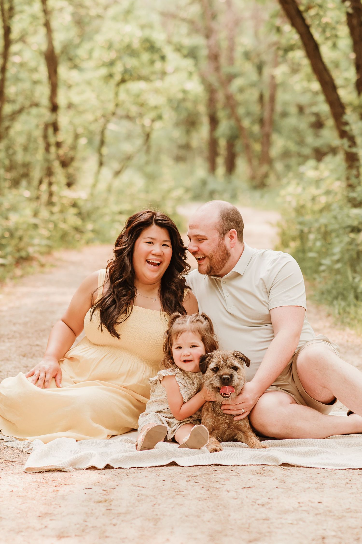Family lying together on blanket in wooded area with their dog while sharing joyful laughter during outdoor photoshoot.