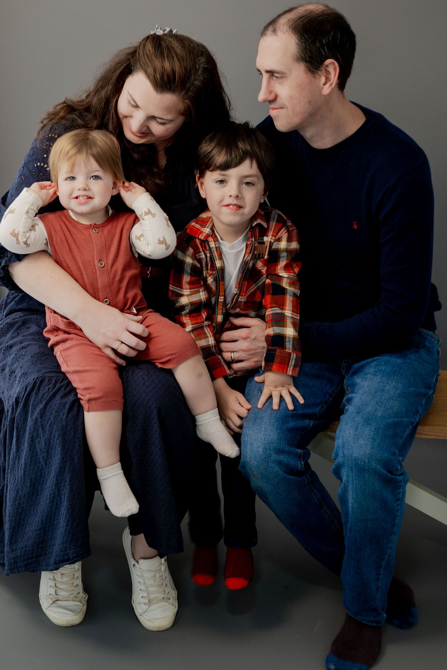 Family portrait with parents in elegant attire holding newborn against white backdrop.