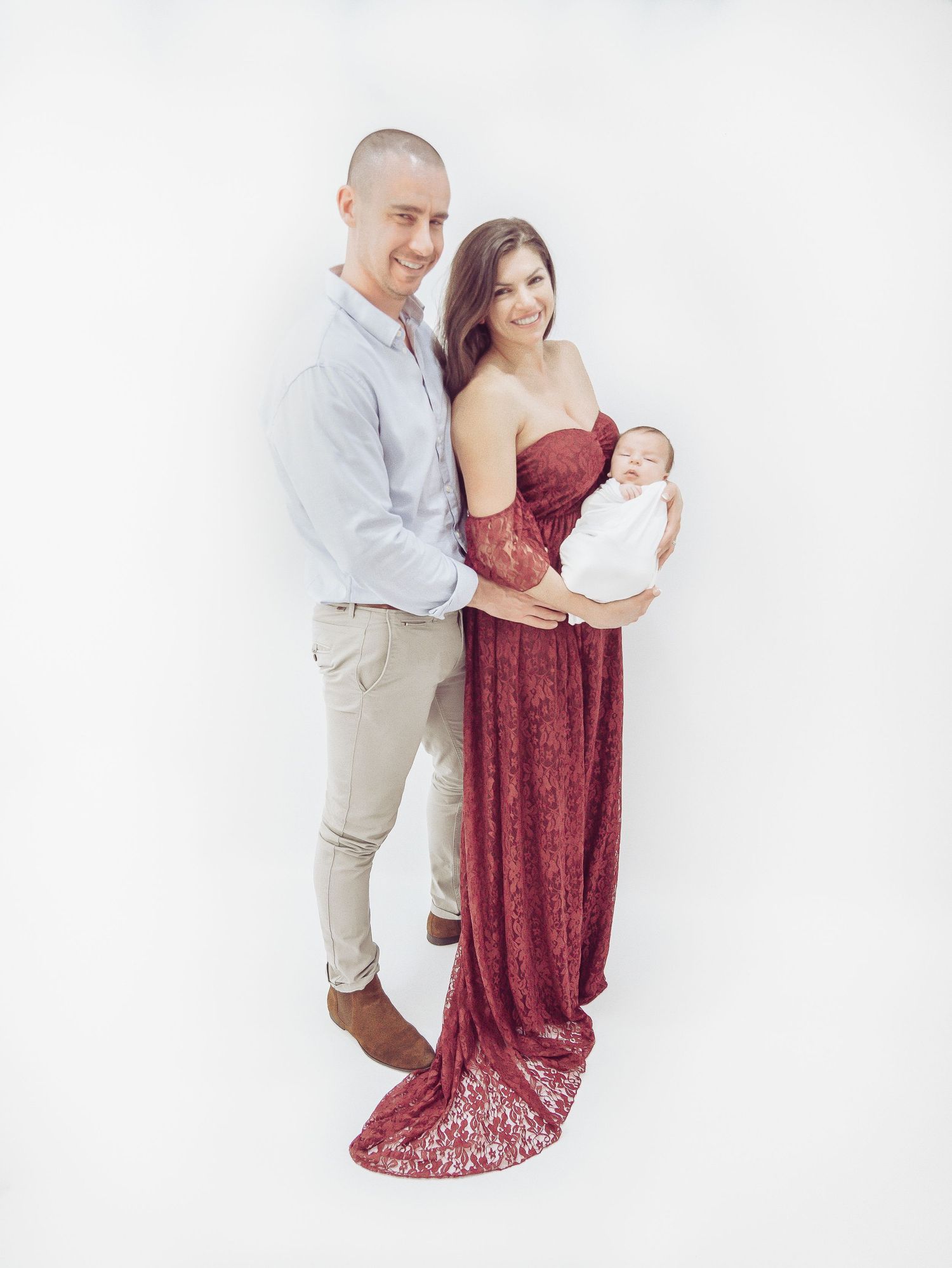 Family portrait with parents in elegant attire holding newborn against white backdrop.