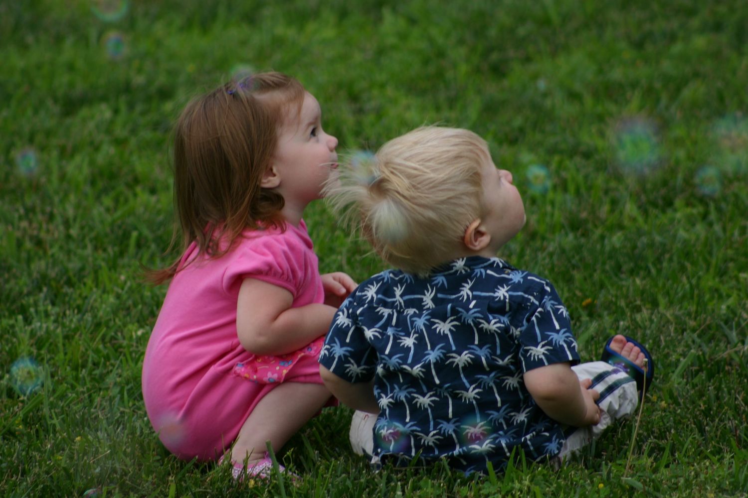 Two young children in pink and blue outfits lay on green grass while playing together outdoors.
