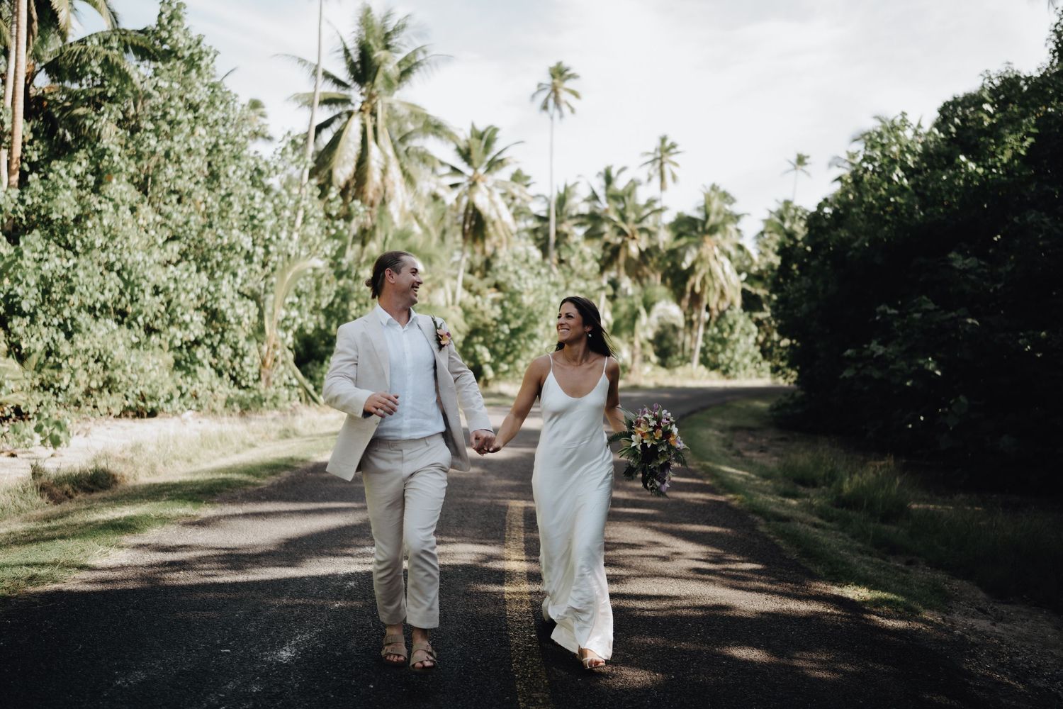 Couple walking hand in hand down a palm tree lined path in tropical wedding attire.