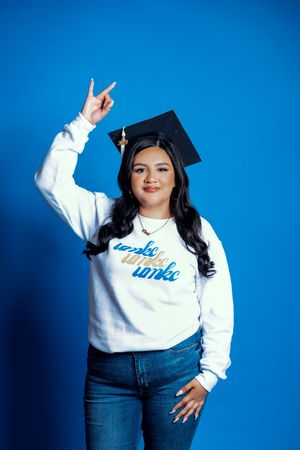 Graduate in white sweatshirt and jeans poses against blue backdrop for celebration photos with graduation cap.