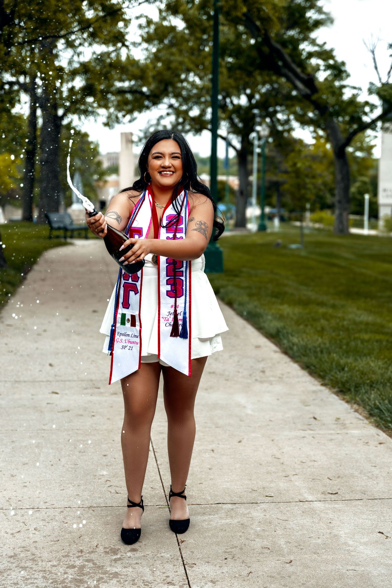Graduate in patriotic stole poses outdoors on campus path during graduation photo session.