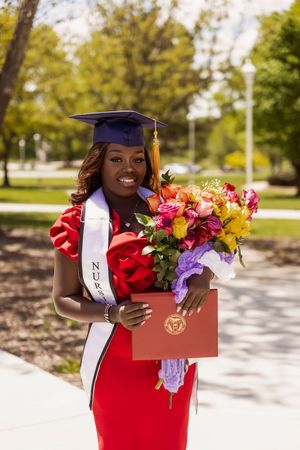 Graduate in red dress holds colorful bouquet of flowers while wearing graduation cap and stole during outdoor ceremony.