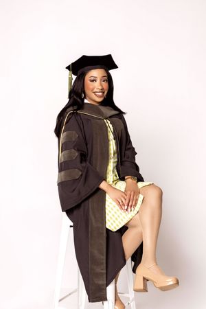 Graduate in black academic regalia poses with gold accessories and nude heels against white backdrop.