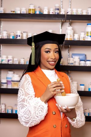 A graduate in an orange vest and white lace sleeves poses with a coffee cup in a pharmacy setting.