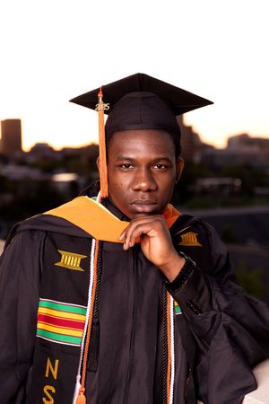 Graduate wearing black academic regalia with colorful kente stole and mortarboard cap poses during sunset.