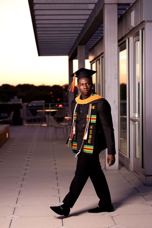 Graduate wearing black academic regalia with colorful kente stole poses in empty hallway at sunset.