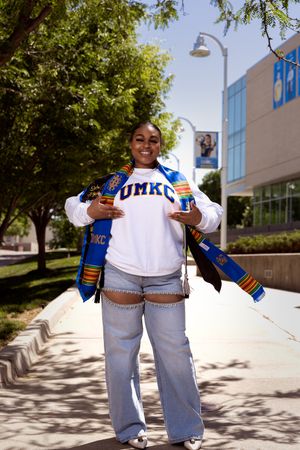 Two people in matching UMKC apparel pose together on a sunny campus sidewalk with trees and buildings visible.