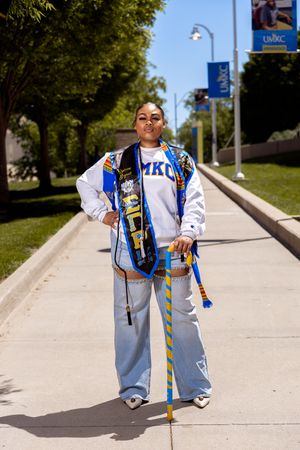 Student wearing white and blue university spirit wear stands on campus sidewalk holding a graduation stole and sash.
