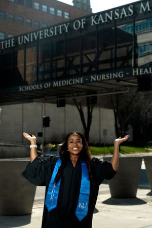 Graduate in blue and black KU regalia celebrates outside university medical building.