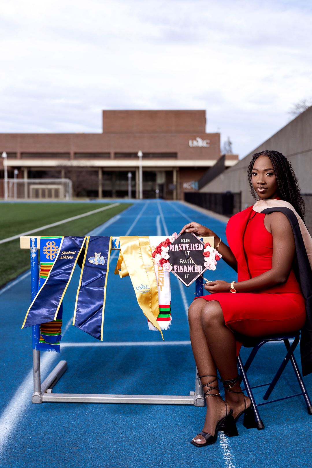 Woman in red dress sits on track holding graduation banner, with academic regalia displayed on rack at UMBC stadium.