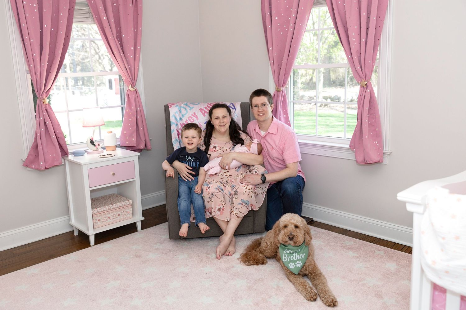 Family of four with newborn and golden retriever dog pose in pink nursery room with curtains and windows.