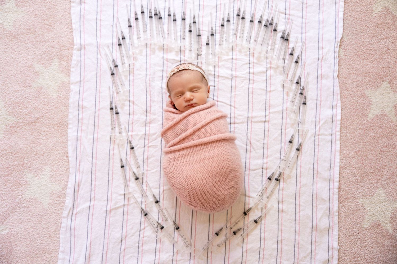 Newborn baby swaddled in pink wrap sleeping inside a heart-shaped arrangement on a soft pink blanket.