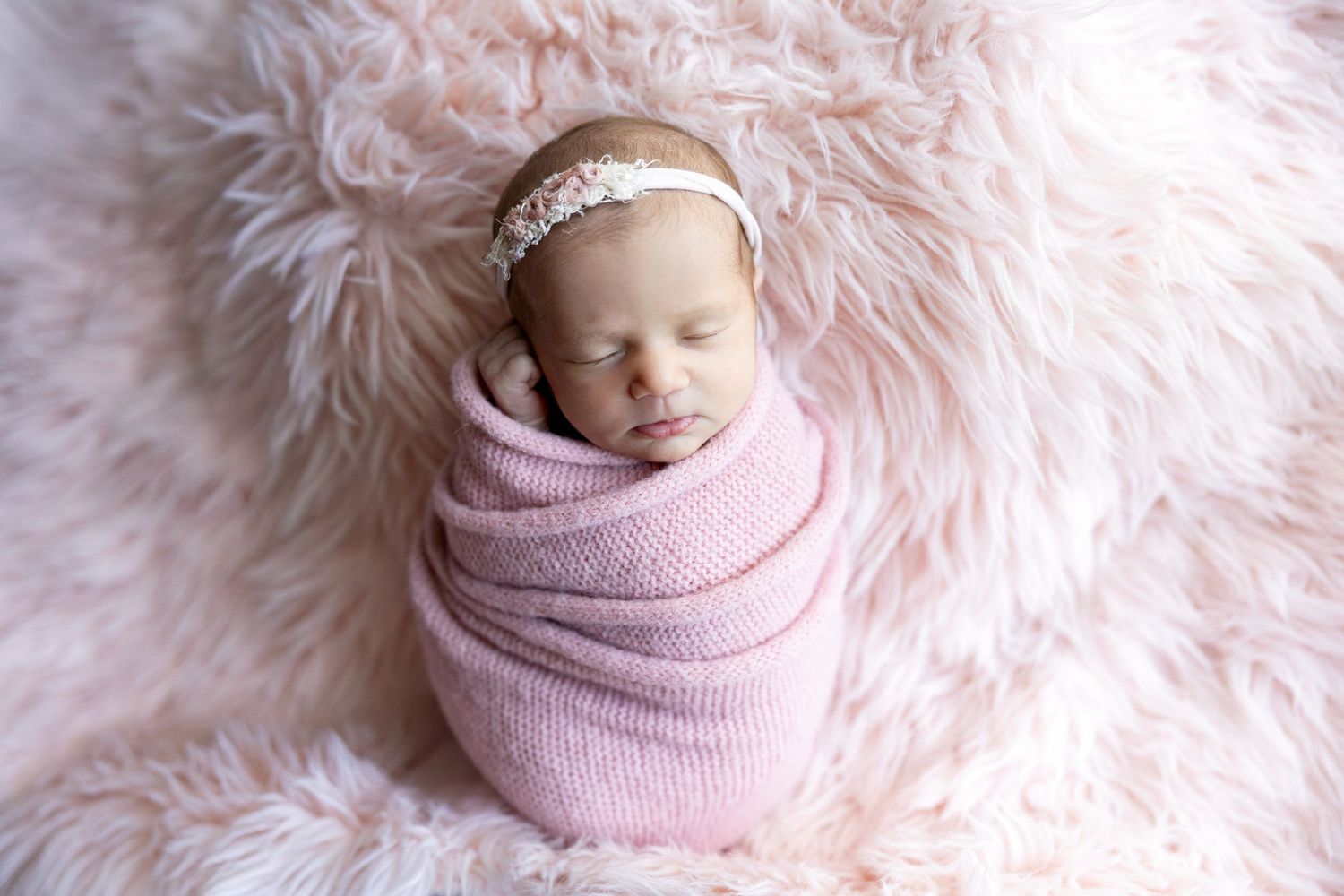 Newborn baby girl swaddled in pink wrap with floral headband, sleeping peacefully on fluffy pink fur rug.