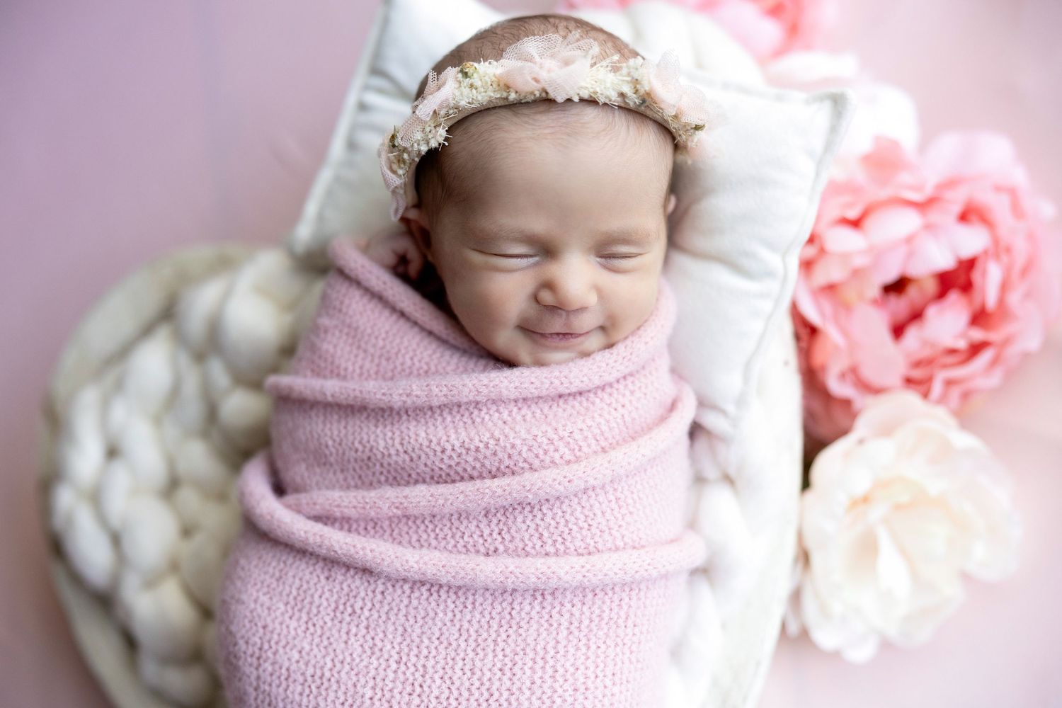 Sleeping newborn baby girl swaddled in pink wrap with floral headband in a basket with flowers.