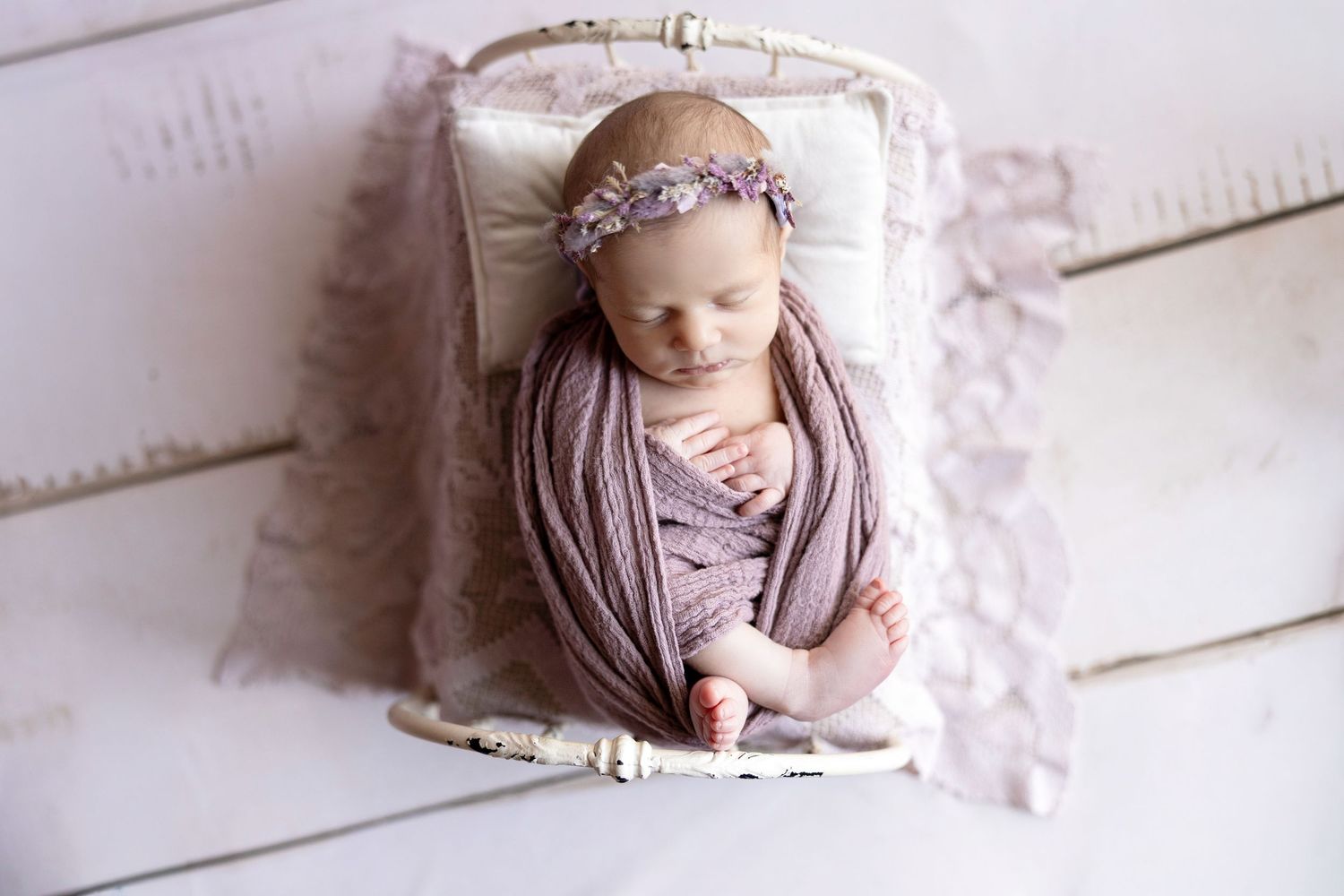 Newborn baby girl wrapped in lavender swaddle with floral headband sleeping peacefully in a white vintage miniature bed.