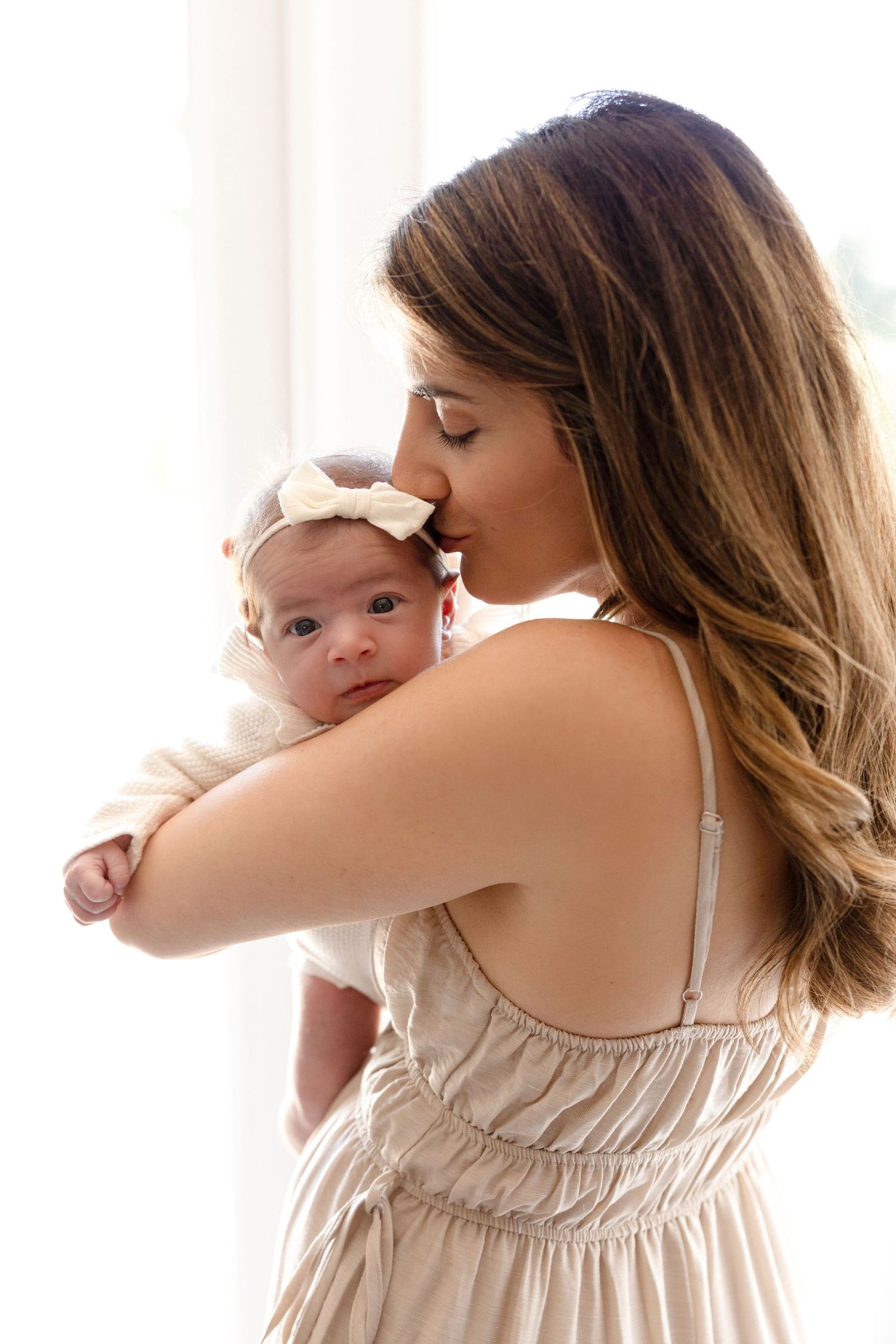Mother in beige dress holds newborn baby near window during lifestyle photoshoot.
