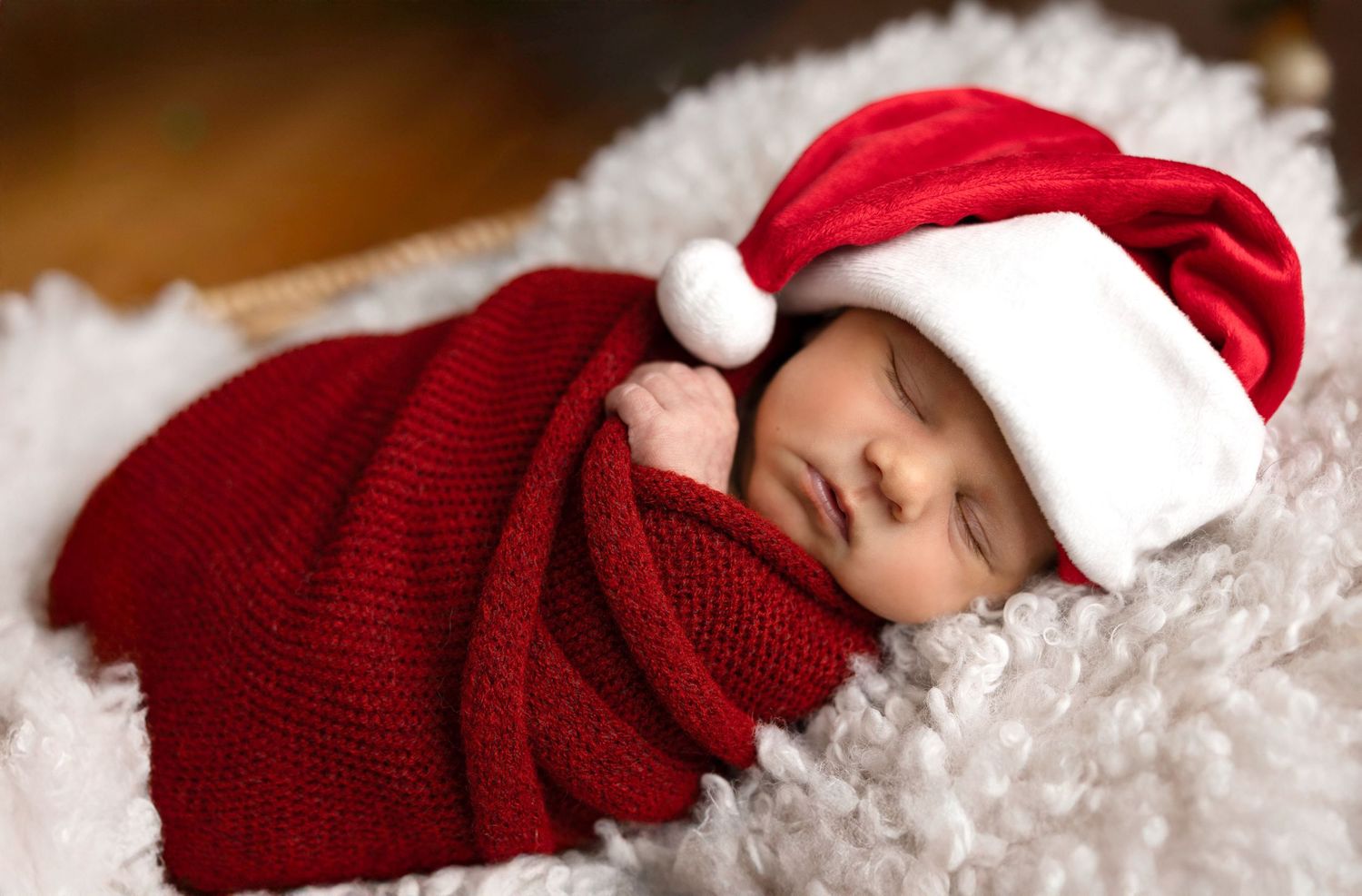 Sleeping newborn wrapped in red knit blanket wearing Santa hat on white fluffy surface.
