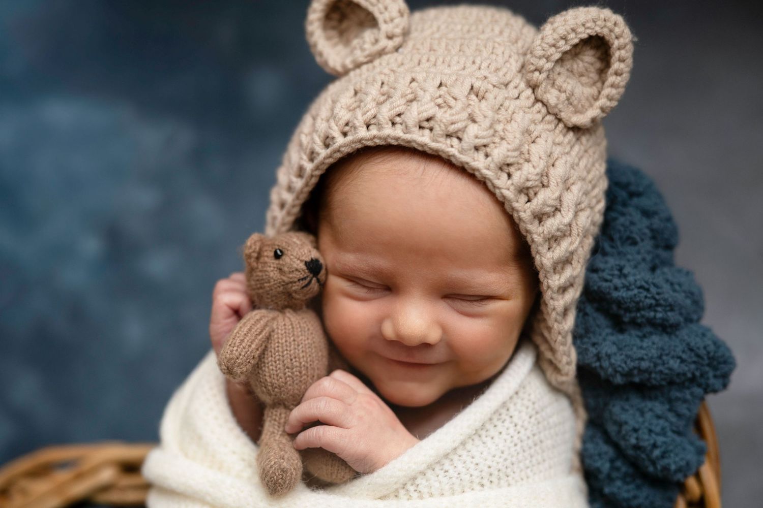 Baby wearing knit bear hat smiling while holding small stuffed toy.
