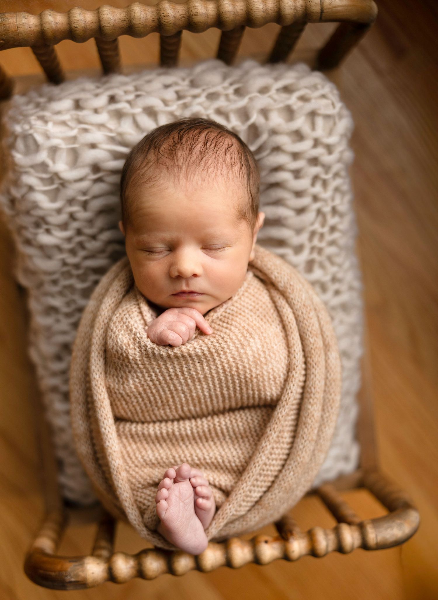 Sleeping newborn wrapped in beige knit blanket on textured chair.