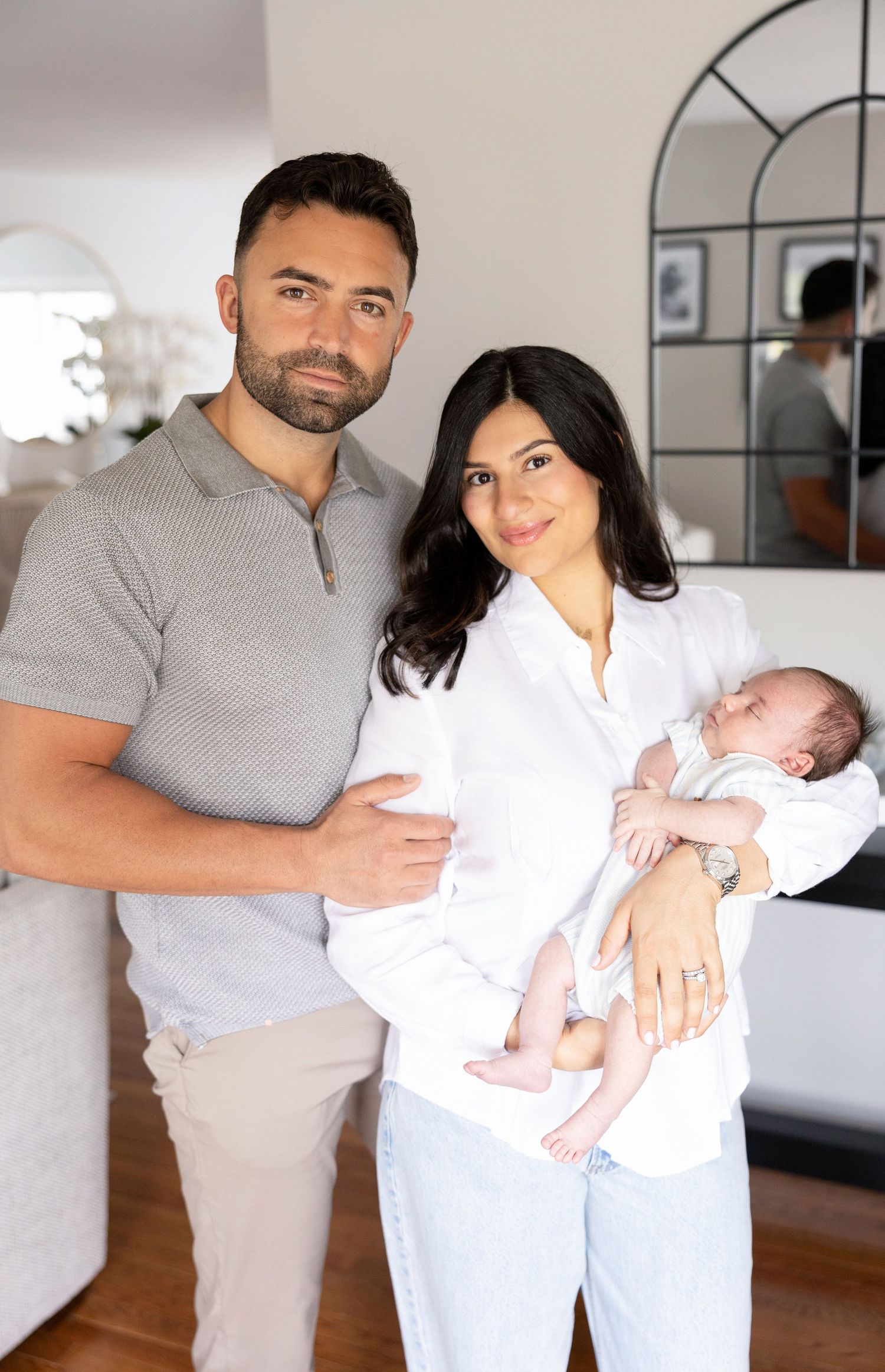 A couple poses indoors holding their newborn baby, the man in a gray polo and the woman in a white shirt and jeans.