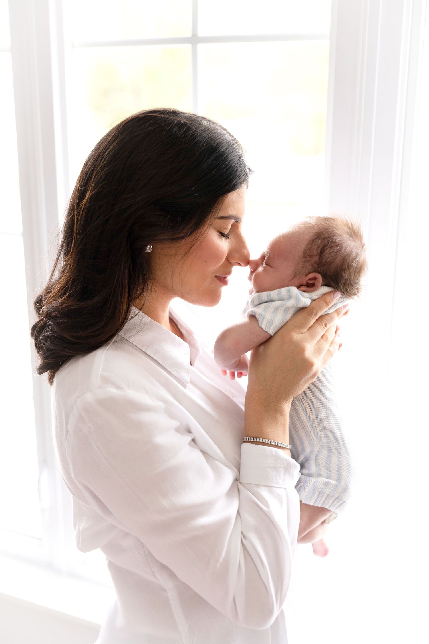 A mother with dark hair tenderly touches noses with her newborn baby near a bright window.