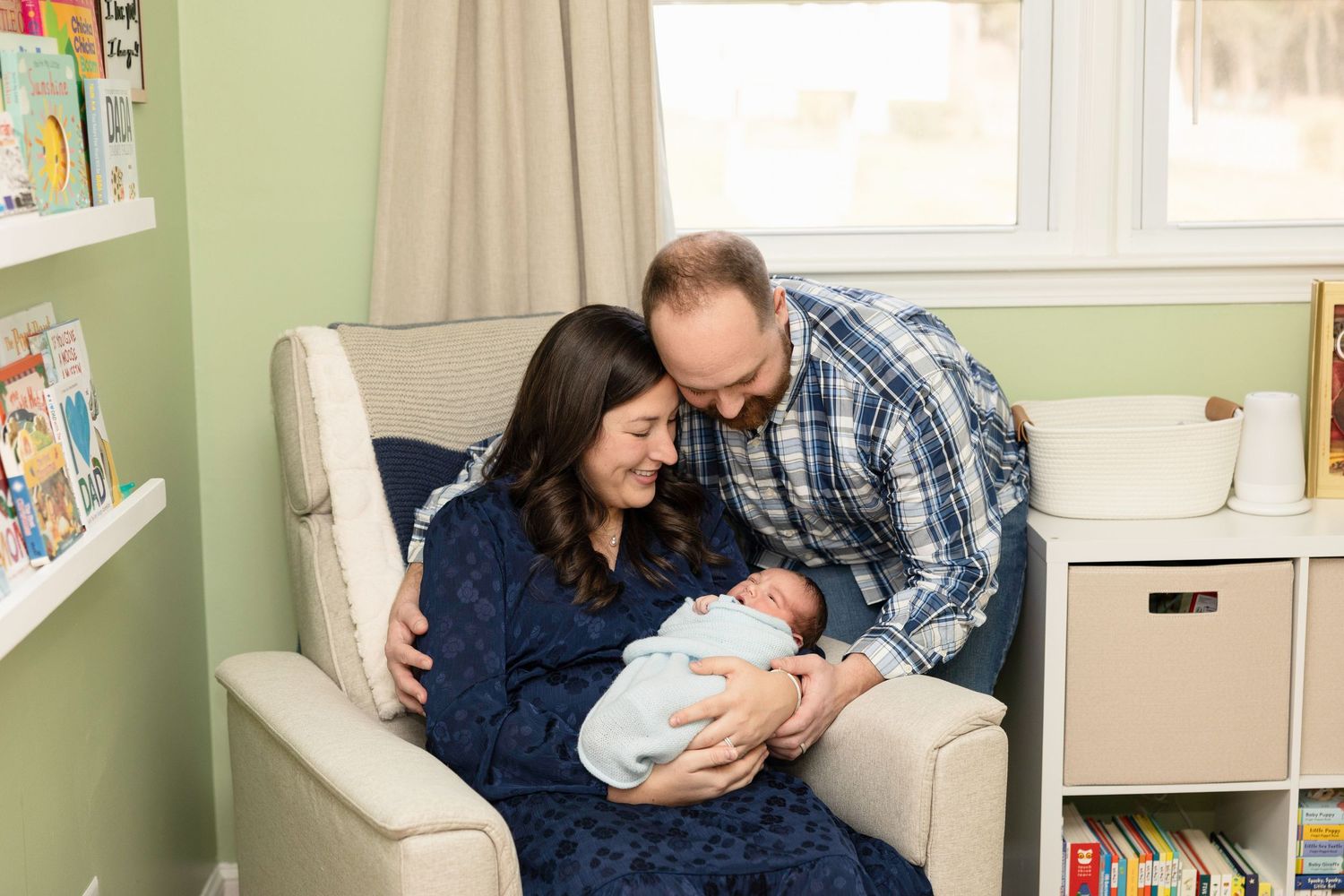 Parents cuddling with newborn in nursery chair surrounded by bookshelves.