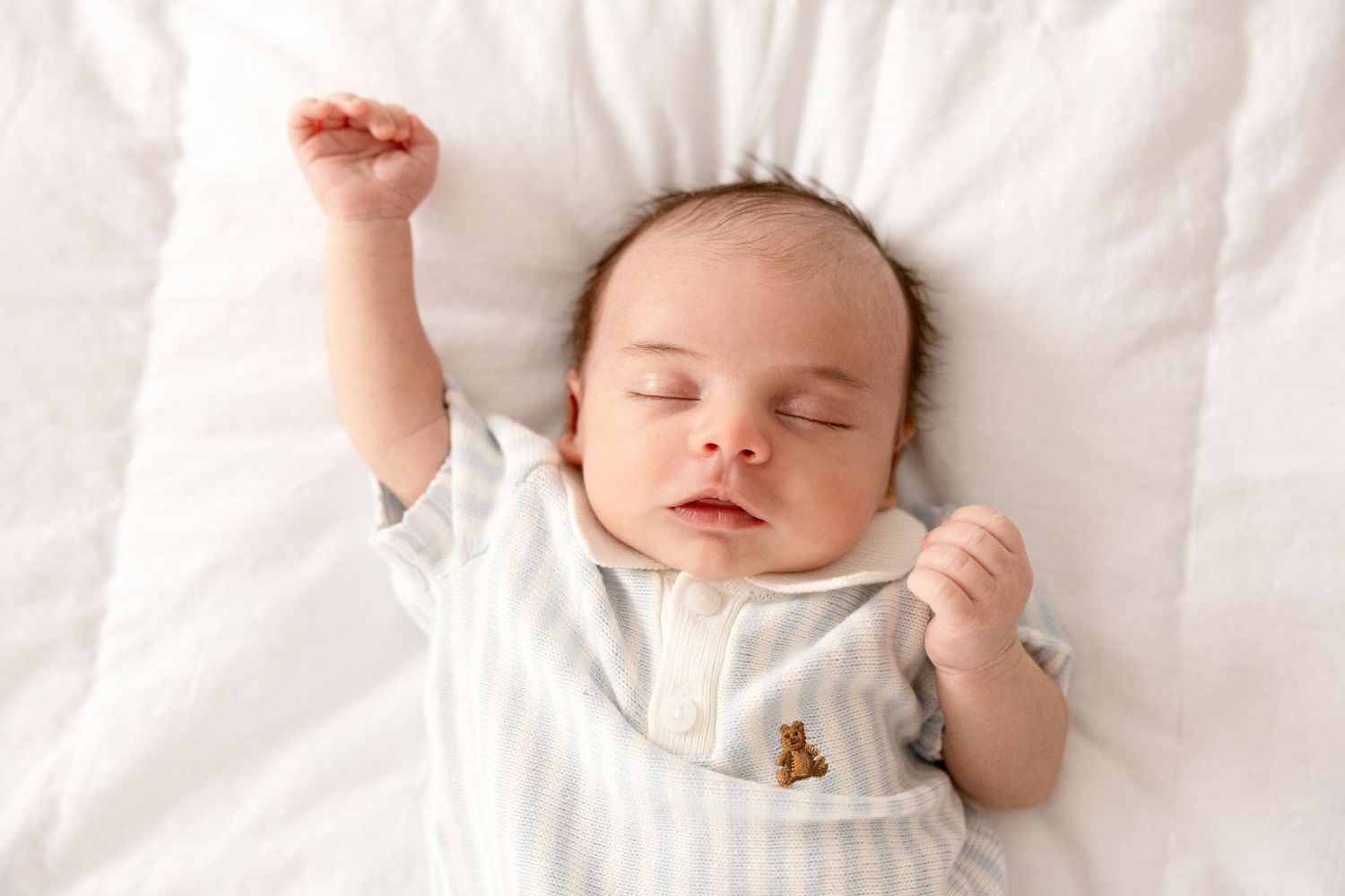 Peaceful newborn baby sleeping on white bedding wearing a white striped onesie with one arm raised above head.